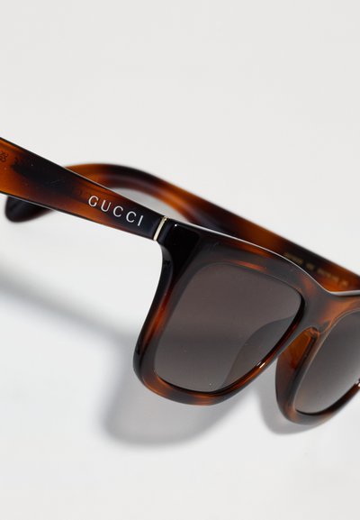 Gucci solbriller med en skildpadde-farvet stel, mørke linser og sølv logo accenter på stængerne, der fremhæver et dristigt, rektangulært design.
