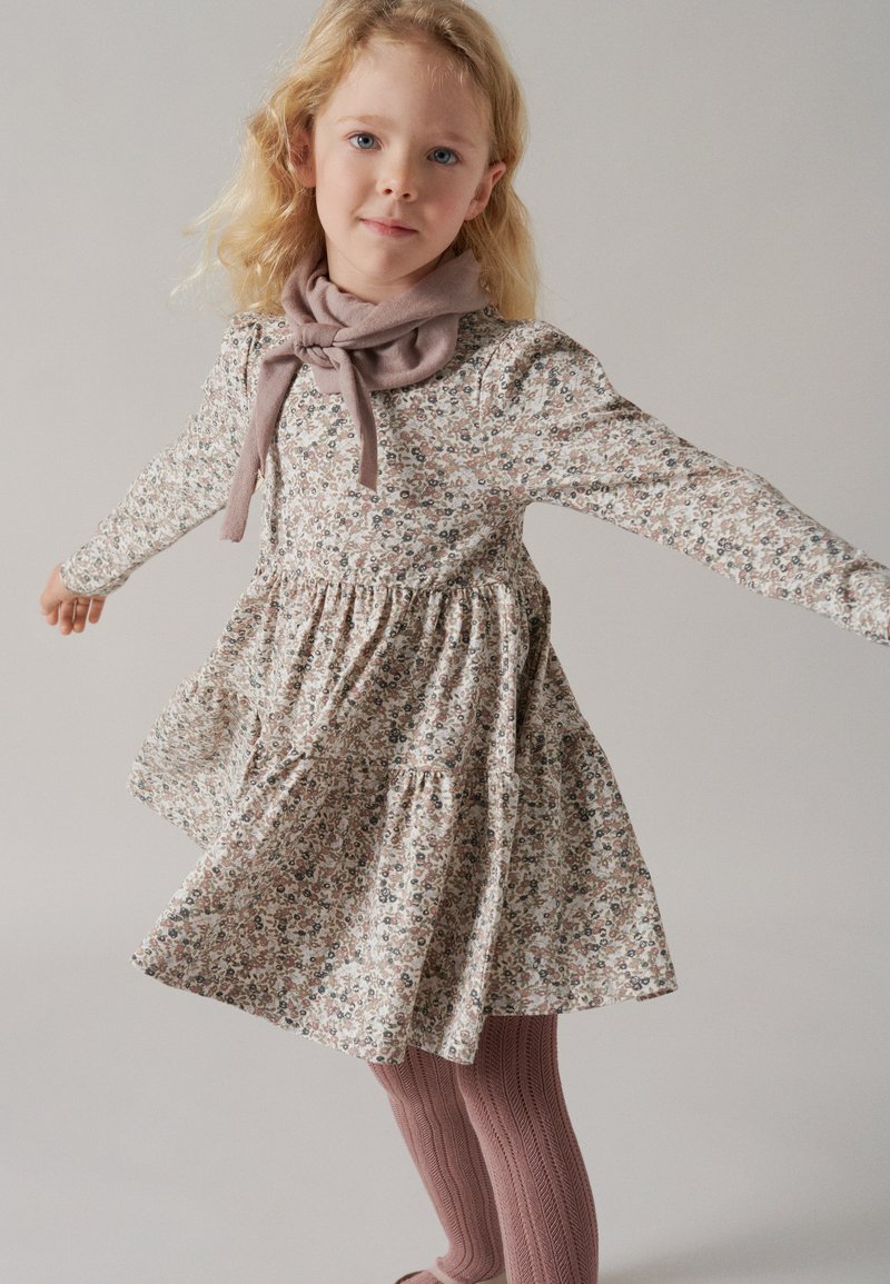 Šaty s kvetinovým vzorom z mäkkého materiálu, s dlhými rukávmi a nazberaným pásom; skombinované s textúrovanými ružovými pančuchami a zodpovedajúcim šálom zviazaným okolo krku.