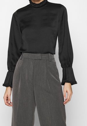 Kobieta ubrana w czarną bluzkę z długim rękawem i falbaniaste mankiety oraz szare, szerokie spodnie z wysokim stanem, stoi na jednolitym tle.