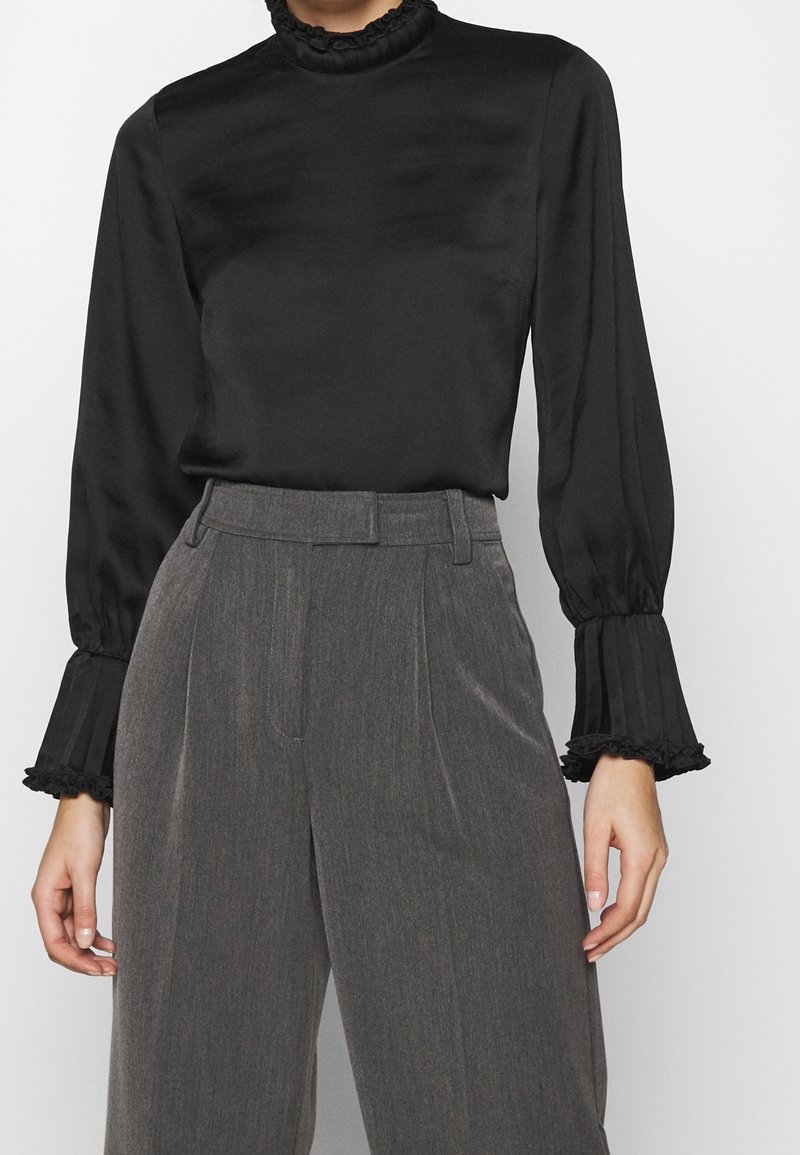 Femme portant un chemisier noir à manches longues avec des poignets à volants et un pantalon gris taille haute à jambes larges, debout devant un fond uni.