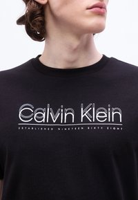 Άτομο που φοράει μαύρο T-shirt με λευκό λογότυπο «Calvin Klein» και κείμενο «Established Nineteen Sixty Eight», γυρισμένο προς τα δεξιά, ορατός ο λαιμός και το πηγούνι.