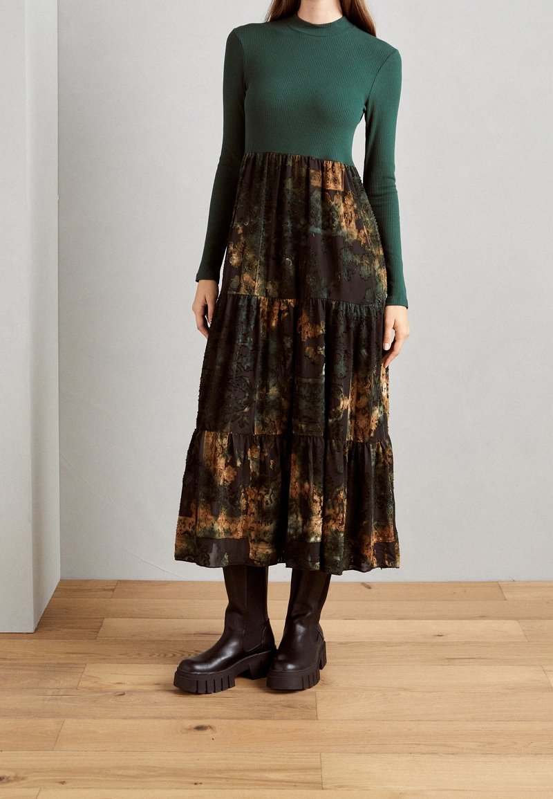 Haut long à manches longues en vert côtelé associé à une jupe noire à motifs floraux en couches ; combiné avec des bottes à talons épais marron sur une surface en bois.