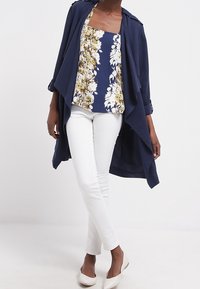 MARINBLÅ ÖPPEN JACKA ÖVER EN BLOMMIG MARINBLÅ OCH VIT CAMISOLE, KOMBINERAD MED VITA SMALA BYXOR OCH NUDFÄRGADE SKOR. Mjuk, lättviktig tyg.