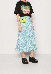 T-shirt graphique noir avec un design de dents colorées, associé à une jupe midi florale bleu clair et des sandales à plateforme noires. Un sac bandoulière vert est visible.