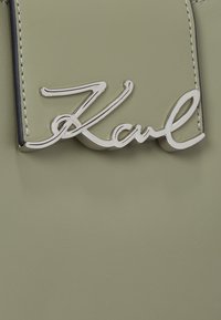 Zelená kožená tkanina s hladkým povrchom, ktorá na seba upozorňuje výrazným strieborným logom "Karl". Jednoduchý dizajn s kontrastným prešitím.