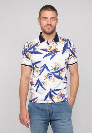 Homme portant un polo blanc avec un motif floral bleu et orange et un jean bleu, debout avec une main dans la poche devant un fond uni.