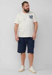 T-shirt en coton crème avec un graphique marine sur la poitrine gauche, associé à un short marine et des baskets blanches. Texture douce, coupe décontractée.
