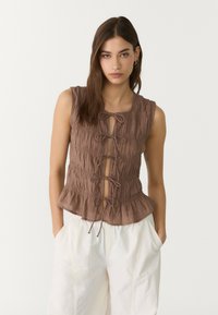 Top péplum sans manches marron en tissu froncé avec un lien de fermeture à l'avant. Présente une texture douce et des détails subtils.
