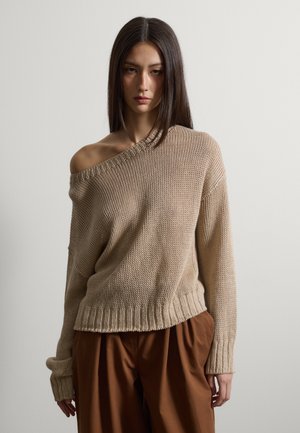 Donna con capelli lunghi e scuri indossa un maglione beige a maglia con scollo a barca e pantaloni marroni larghi, in piedi davanti a uno sfondo chiaro semplice.
