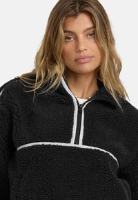 Schwarzer Fleece-Pullover mit weißen Akzenten, halbem Reißverschluss und strukturiertem Material. Bietet eine lässige Passform und ausgeprägte Nahtdetails.