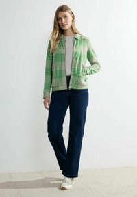 Chaqueta a cuadros verde y beige con cierre frontal, dos bolsillos laterales, combinada con jeans azul oscuro y zapatillas de color claro.