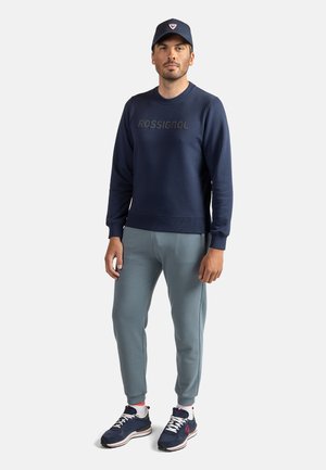 Marineblå sweatshirt med "ROSSIGNOL" i hævet skrift, lysegrå sweatpants og matchende marineblå cap. Sportssko med røde og hvide detaljer.