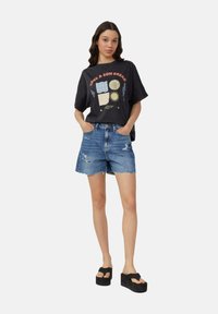 T-shirt graphique noir décontracté avec un motif coloré de soleil et de vagues, associé à un short en denim bleu taille haute avec des bords effilochés. Sandales à plateforme noires.