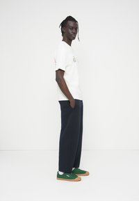 Νεαρός άνδρας στέκεται πλαγίως με τα χέρια στις τσέπες, φοράει λευκό μπλουζάκι με γραφικό σχέδιο, ναυτικό μπλε παντελόνι και πράσινα αθλητικά παπούτσια με σόλες από καουτσούκ.