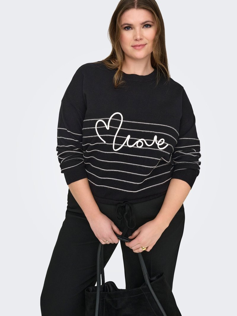 Femme portant un pull noir à rayures blanches avec le texte "love", tenant un sac noir, debout devant un fond clair uni.