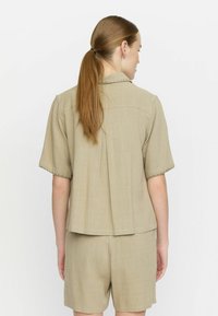 Light brown short-sleeve shirt with a collar, textured fabric, and frayed edges, paired with matching shorts, seen from the back.