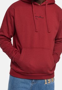 Dunkelroter Kapuzenpullover mit Fronttasche, Kordelzug an der Kapuze und dezentem schwarzem Logo auf der Brust. Hergestellt aus weichem, strukturiertem Stoff.