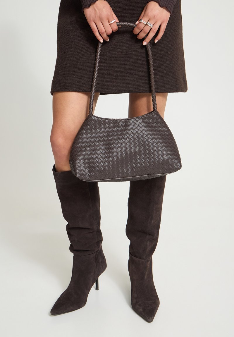 Femme tenant un sac à main tissé marron foncé, portant une mini-jupe noire et des bottes hautes à talons en daim marron foncé jusqu'au genou.