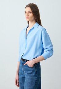 Camicia azzurra leggera con colletto, texture morbida e vestibilità rilassata, abbinata a jeans blu a vita alta.