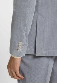 Manica di un blazer in velluto a coste grigio chiaro con quattro bottoni beige, caratterizzato da una texture a coste verticali e una vestibilità su misura.