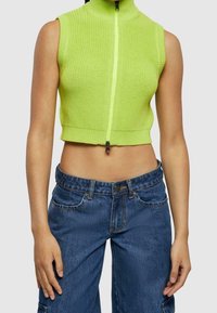 Top de punto corto en verde neón con cuello alto y cremallera frontal, combinado con pantalones vaqueros de pierna ancha en azul denim que cuentan con bolsillos delanteros.