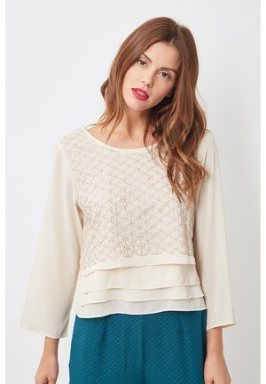 Blusa crema con un panel frontal estampado, dobladillo en capas y mangas fluidas. Tejido suave con detalles de bordado geométrico.