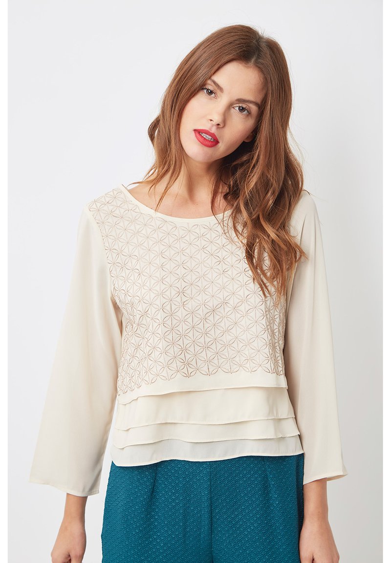Blusa crema con un panel frontal estampado, dobladillo en capas y mangas fluidas. Tejido suave con detalles de bordado geométrico.