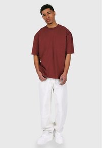 Bordeaux oversized t-shirt med korte ærmer, parret med hvide slidte jeans og hvide sneakers, stående på en neutral baggrund.