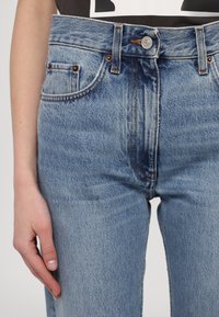 Šviesiai mėlyni džinsai su aukštu liemeniu, papuošti variniais nagais, siuvinėjimu ir užsegimu mygtuku. Matomos priekinės kišenės.