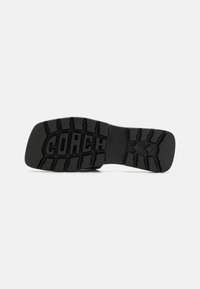 Talpa de pantof negru din cauciuc, cu un grip texturat, având cuvântul "COACH" afisat proeminent, înconjurat de un model de profil și detalii de design.