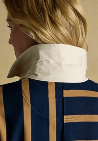 Blonde woman with navy and camel striped jacket and raised cream collar embroidered with "Joules" on the back.
