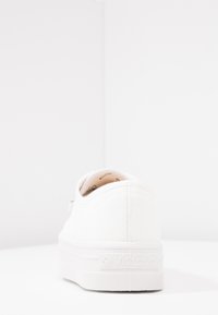 Baskets en toile blanches avec une surface texturée, semelle plate et détail du logo sur le talon. Design minimaliste et plateforme légèrement surélevée.