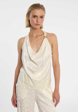 Femme portant un haut licou en satin couleur crème avec un détail anneau en or et un pantalon assorti à motif animalier subtil, debout devant un fond blanc.