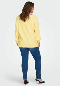 Geltonas megztinis su iškiliais raštais ir pūstomis rankovėmis, derinamas su mėlynais siaurais džinsais ir juodais aukštakulniais sandalais, matomas iš nugaros.