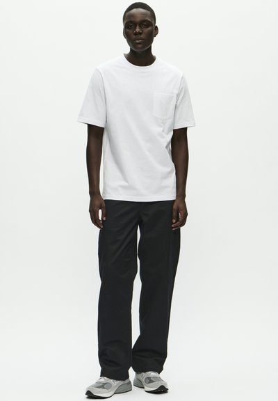 Mann stående vendt mot kameraet, iført en hvit kortermet T-skjorte med lomme, svarte bukser og grå joggesko foran en hvit bakgrunn.