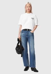 Woman with straight blonde hair wearing white logo t-shirt, blue jeans, black belt, black ankle boots, holding black handbag with wide strap.