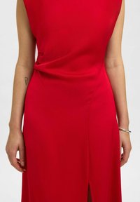 Frau trägt ein ärmelloses rotes Kleid mit leichtem Drapé an der Taille, kleinem Schlitz vorne, sichtbarem Tattoo am linken Arm und silbernem Armband am rechten Handgelenk.