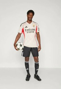 Maillot à domicile du Benfica avec un maillot beige clair agrémenté d'accents rouges, un short noir et des chaussures noires. Tenant un ballon de football noir et blanc.