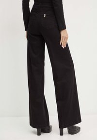 Jeans neri a vita alta e gamba larga con una texture liscia, dotati di due tasche posteriori e dettagli in metallo argentato. Indossati con tacchi a piattaforma neri.