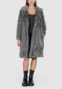 Cappotto in pelliccia sintetica grigia con colletto, chiusura a doppiopetto e texture morbida. Indossato sopra un vestito aderente scuro e stivali neri.