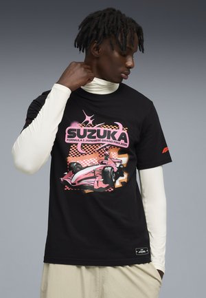 Ung mand iført sort Suzuka 2026 T-shirt med racertema over hvide lange ærmer, kigger nedad, grå baggrund.