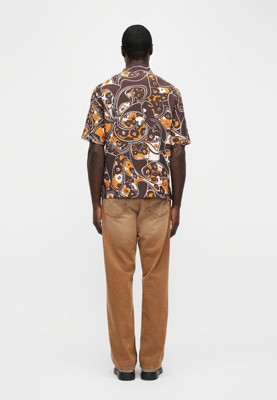 Άνδρας που στέκεται πλάτη, φορώντας καφέ κοντομάνικο πουκάμισο με πορτοκαλί ανθικό μοτίβο, καφέ παντελόνια και μαύρα παπούτσια σε λευκό φόντο.