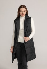 Zwarte gewatteerde vest met een hoge kraag, voorzien van een knoopsluiting en zijzakken. Draagt over een witte geribde turtleneck en donkere jeans.