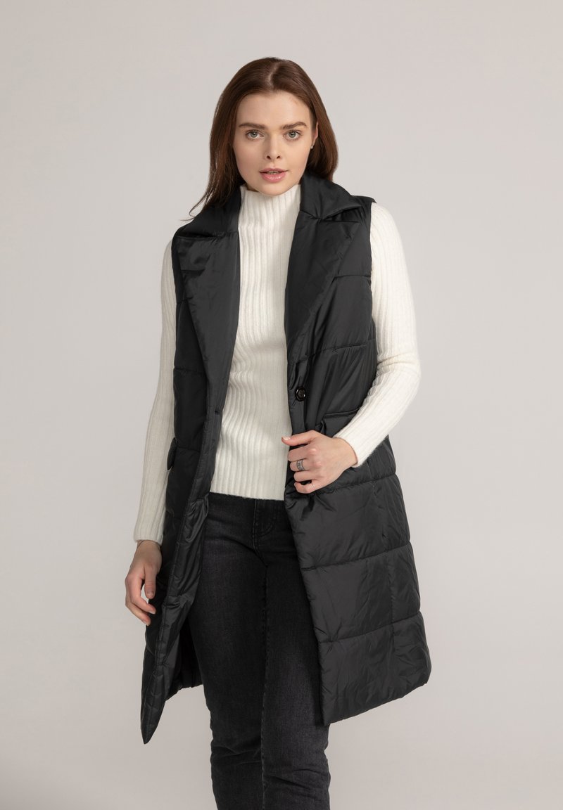 Zwarte gewatteerde vest met een hoge kraag, voorzien van een knoopsluiting en zijzakken. Draagt over een witte geribde turtleneck en donkere jeans.