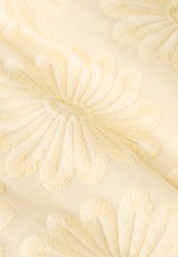 Tissu crème avec des motifs floraux brodés en jaune pâle, présentant de grands designs texturés en forme de pétales créant une apparence douce et élégante.