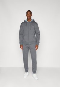 Grijze rits hoodie met trekkoorden en voorzakken, gecombineerd met bijpassende trainingsbroek. Beide gemaakt van zachte stof, aangevuld met witte sneakers.