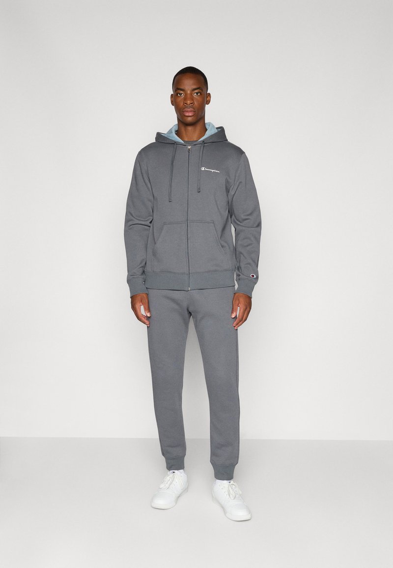 Grijze rits hoodie met trekkoorden en voorzakken, gecombineerd met bijpassende trainingsbroek. Beide gemaakt van zachte stof, aangevuld met witte sneakers.