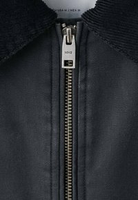 Čierna bunda so zamatovým golierom a kovovým zipsom. Zips má obdĺžnikový ťahák a textúrovaná tkanina je jasne viditeľná.