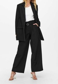 Nainen, jolla on päällään musta ylisuuri bleiseri, vyöllä varustetut korkeavyötäröiset leveälahkeiset mustat housut, valkoinen paita ja läpinäkyvillä remmeillä varustetut korkokengät.