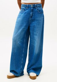 Weite blaue Denim-Jeans mit einem verwaschenen Look, Taschen vorne und einem Knopfverschluss. Kombiniert mit hellen Sneakers.
